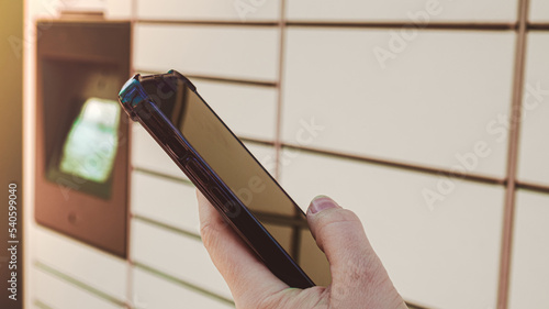 Female hand holding mobile phone next to Parcel locker Collecting parcel from shopping locker. Woman Skans QR Code on Mobile phone Self-service Locker Cell with Bar code Reader Modern Shipping and photo