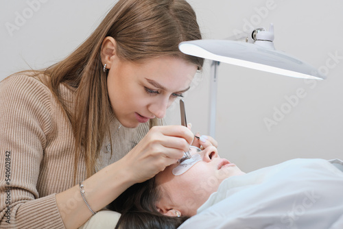 young woman beautician holding tweezers with false lash during lash extension procedure with client. beauty care salon, professional face care.