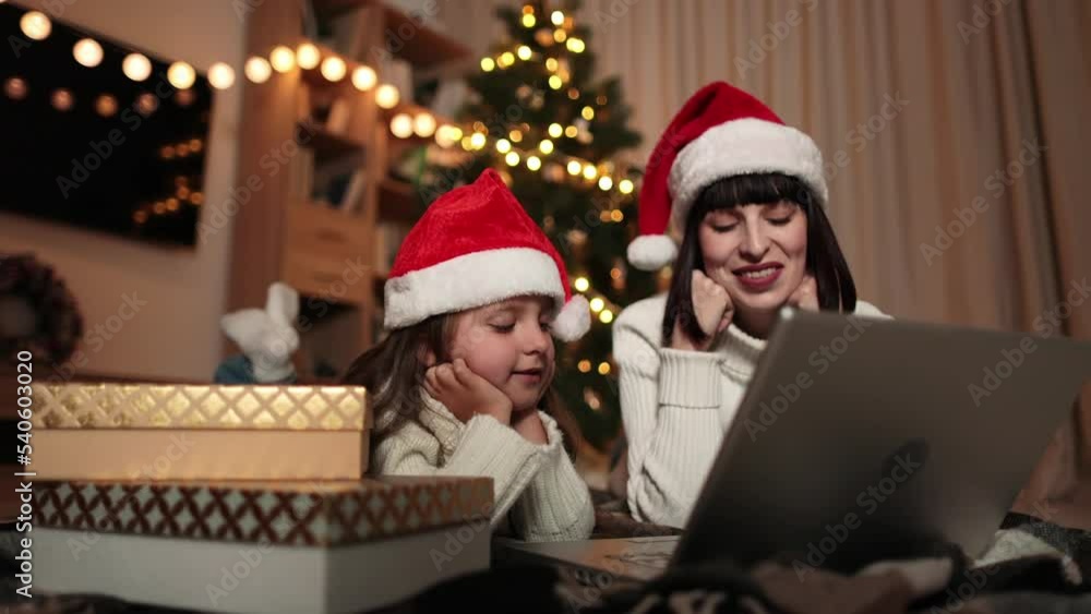 Indoor shot of beautiful happy young woman and her girl shopping online on laptop in cozy Christmas interior. Mother on the floor next the Christmas tree and daughter lying near her, shopping gifts. Indoor shot of beautiful happy young woman and her girl shopping online on laptop in cozy Christmas interior. Mother on the floor next the Christmas tree and daughter lying near her, shopping gifts.
