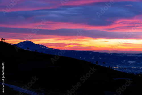 A beautiful colorful epic reddish orange sunrise sky with mountain range and beautiful city lights - Magelang Regency looking out from slope of Mount Sumbing