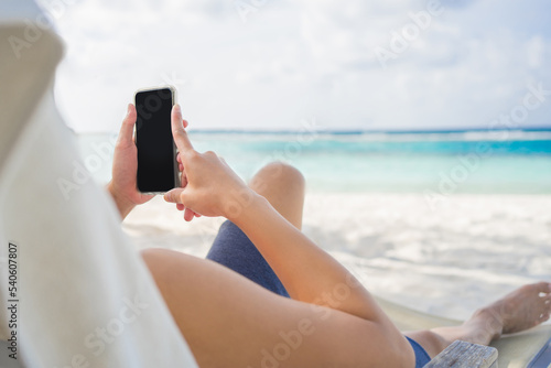 Young Asian man relaxing and lying on beach chair on tropical island beach using smartphone with internet for working or online shopping in summer holiday vacation trip.