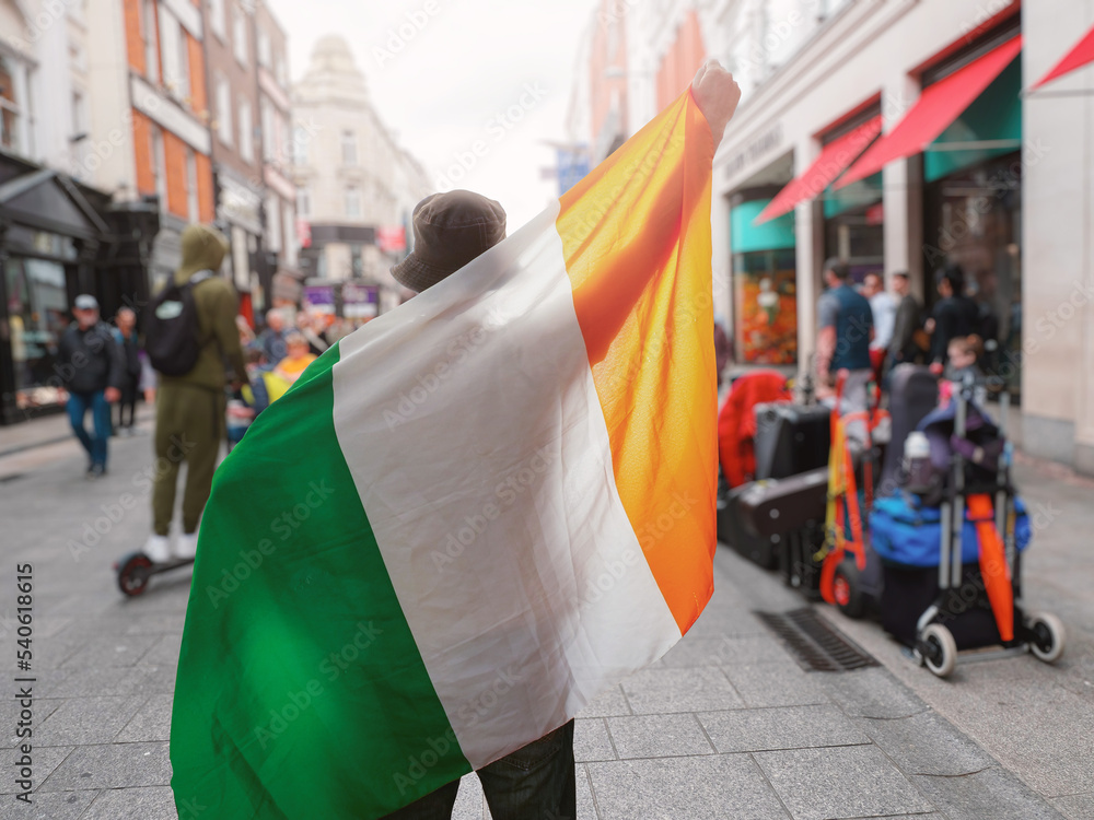 Fototapeta premium Man holding National flag of Ireland in focus. Grafton street of Dublin city out of focus in the background. Support or protest concept. Sun flare. Male with Irish flag.