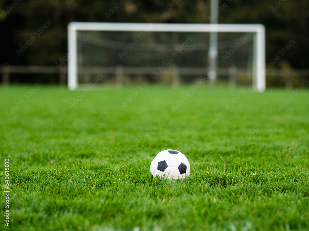 Classic soccer of football ball on the dark green grass field in focus. Goal post out of focus in the background. World wide popular sport activity. Classic soccer of football ball on the dark green grass field in focus. Goal post out of focus in the background. World wide popular sport activity.