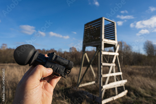 On the hunt, a thermal imaging monocular in hand and a hunting tower in nature.