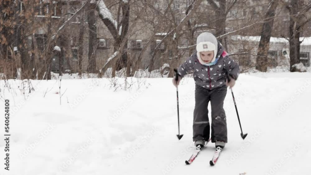 A little girl learns to ski downhill in winter on snow, a focused face. Pushes off with sticks, slides down on bent legs, awkward movements of an inexperienced skier. Snow is falling. A little girl learns to ski downhill in winter on snow, a focused face. Pushes off with sticks, slides down on bent legs, awkward movements of an inexperienced skier. Snow is falling.