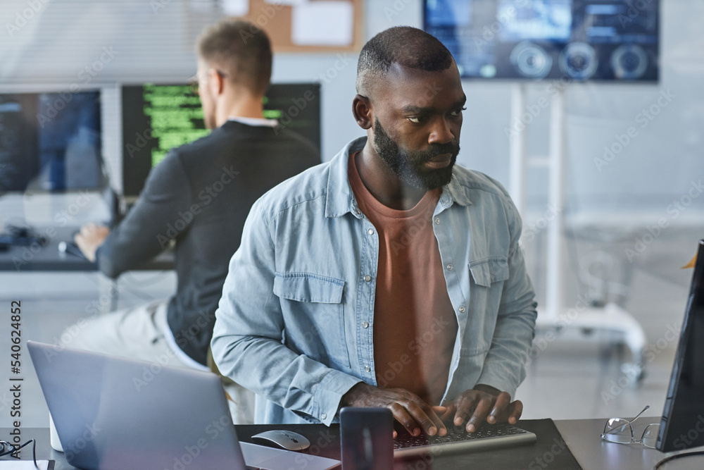Fototapeta premium Portrait of young black man using computer while programming software in IT company office behind glass wall