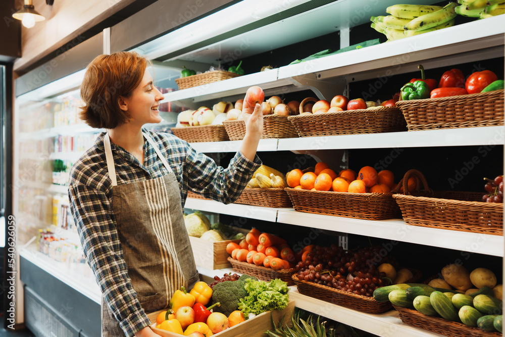 Happy Caucasian young adult employee is looking at fruit shelves and preparing to add an apple to the shelf. Saleswoman with an apron is holding an apple and managing the stock of fruit on shelves.