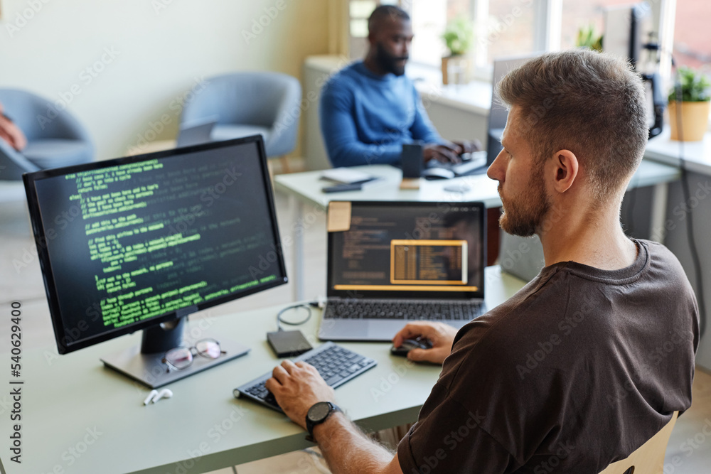 Portrait of software engineer writing code at workplace in office with multiple devices