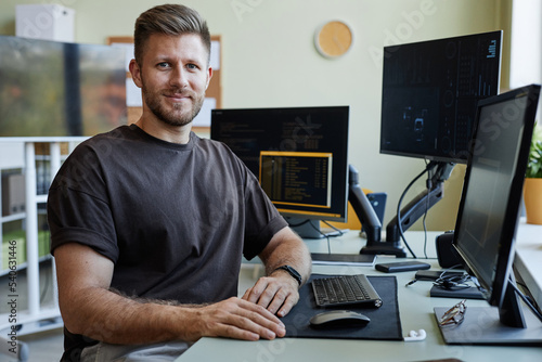Portrait of smiling software developer looking at camera while sitting at desk with computers