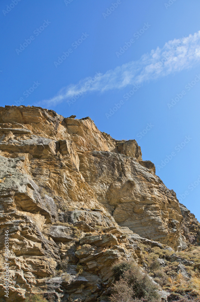 A dry shist rock cliff face under a blue sky with cloud line on the Lake Dunstan Cycle Trail .