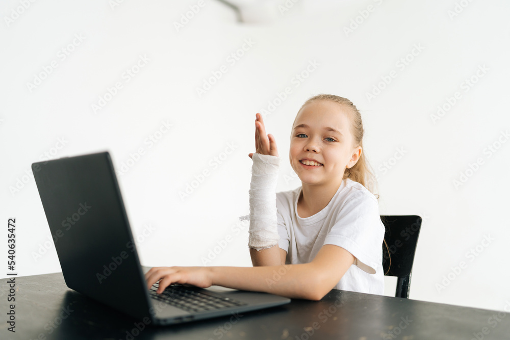 Cheerful blonde little girl raising broken hand wrapped in white plaster bandage sitting at table with laptop smiling looking at camera in light room. Concept of child insurance and healthcare. Cheerful blonde little girl raising broken hand wrapped in white plaster bandage sitting at table with laptop smiling looking at camera in light room. Concept of child insurance and healthcare.