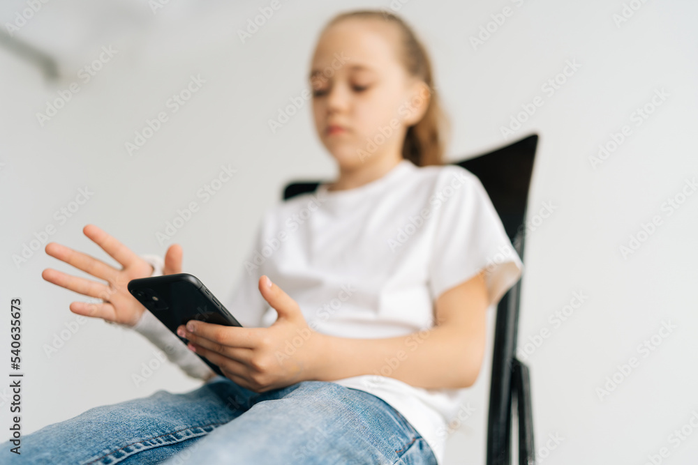 Close-up low-angle view of blonde little girl with broken arm wrapped in white plaster bandage using typing smartphone sitting at chair in light room. Concept of child insurance and healthcare. Close-up low-angle view of blonde little girl with broken arm wrapped in white plaster bandage using typing smartphone sitting at chair in light room. Concept of child insurance and healthcare.