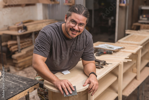 A man smiles at the camera while sanding the edges of a newly built shoe bench at his furniture workshop.