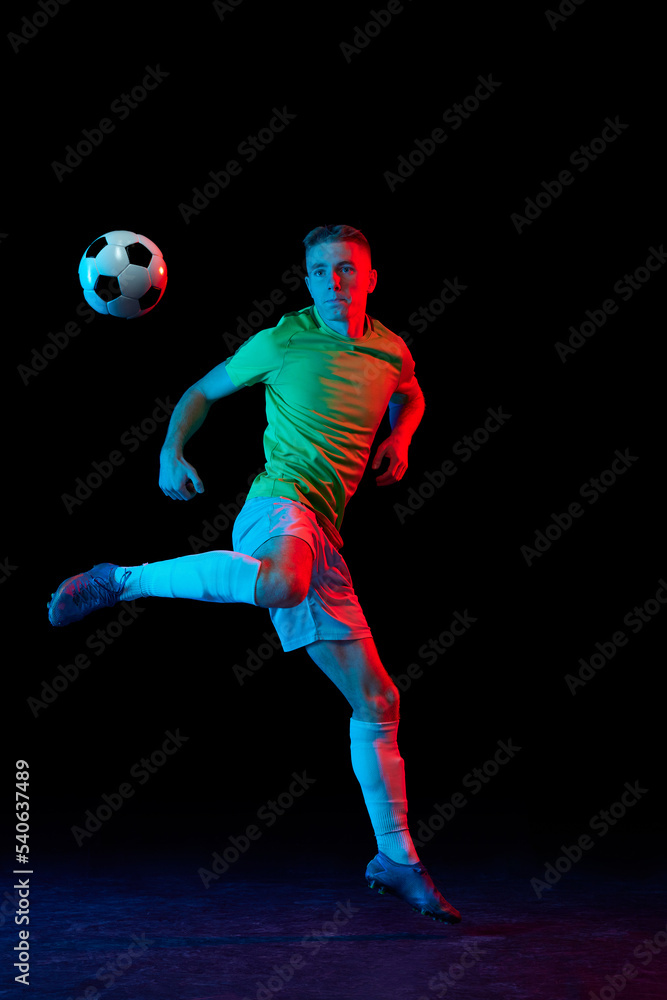 Dynamic shot of young active football player in action isolated on dark background in neon light. Concept of sport, goals, competition, achievements.
