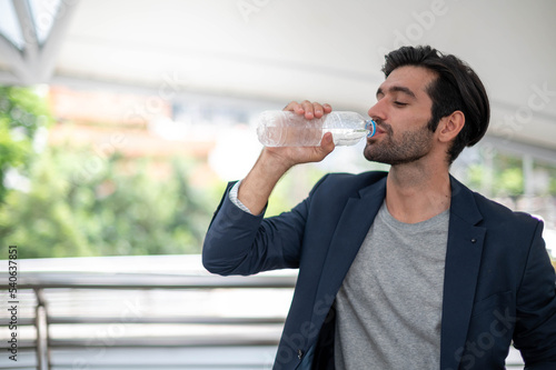 The businessman is drinking water at his watch and wearing a deep blue suit while he resting outside.