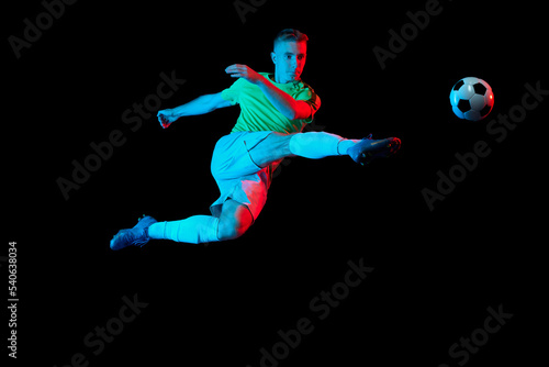 Sport in action. One man professional soccer player training with football ball isolated on dark background in neon light filter. Sport, speed, power and energy