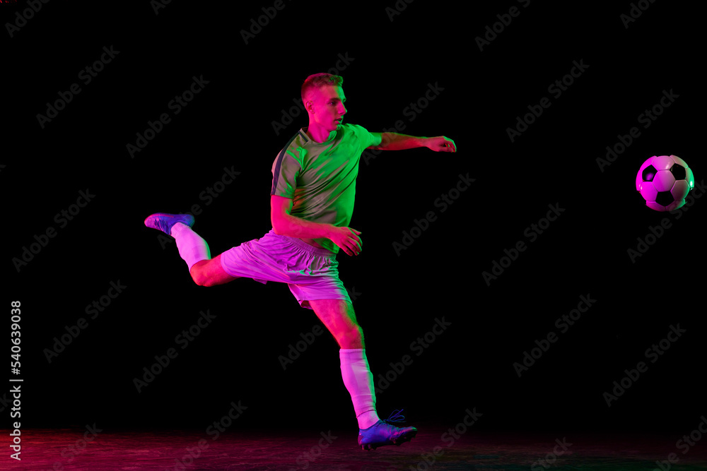 Young professional male football soccer player in motion isolated on dark background in neon light. Concept of sport, goals, competition, league, ad.