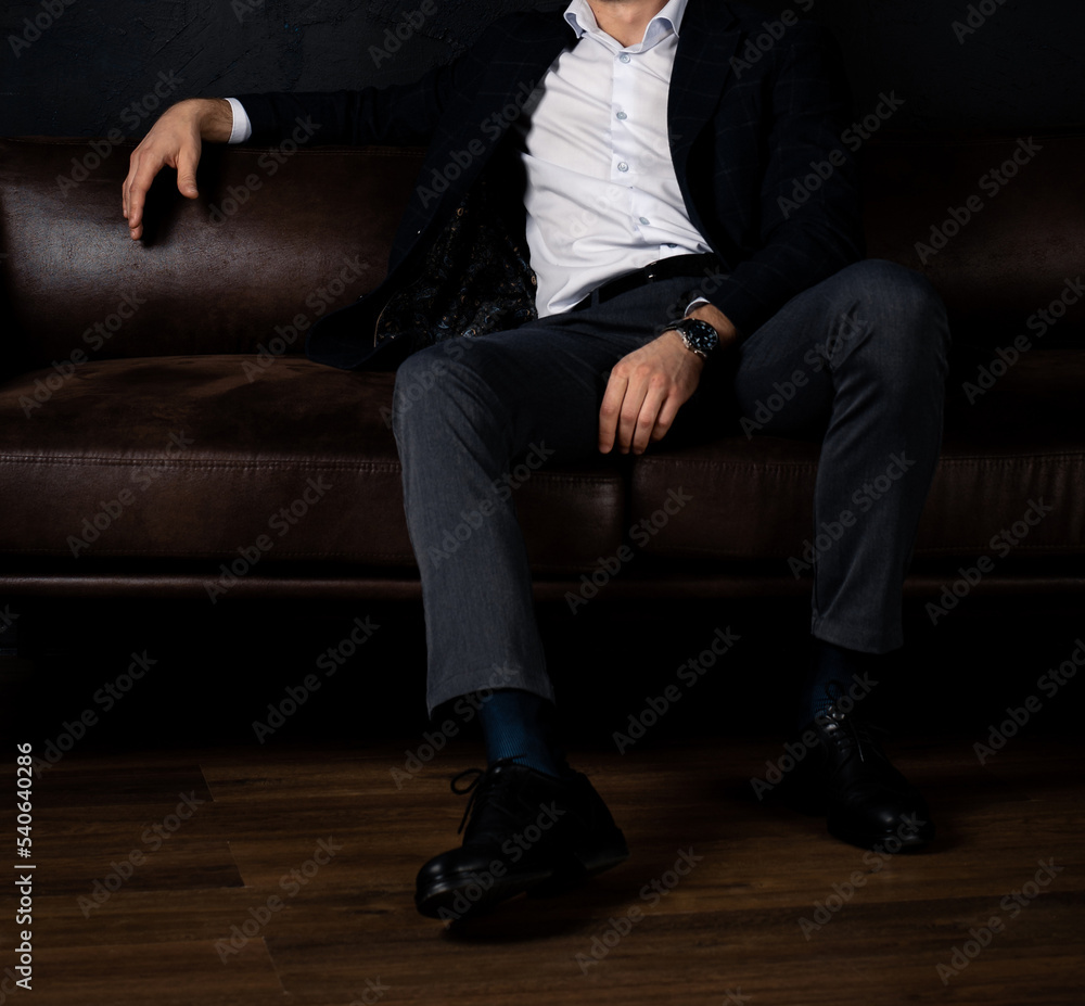 An unrecognizable man in stylish clothes sits relaxed on the sofa in the dark. An unrecognizable man in stylish clothes sits relaxed on the sofa in the dark.