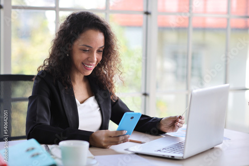 Attractive professional latin female employee worker sitting, using laptop computer with paperwork at home workplace. Businesswoman using smart mobile phone while holding credit card.