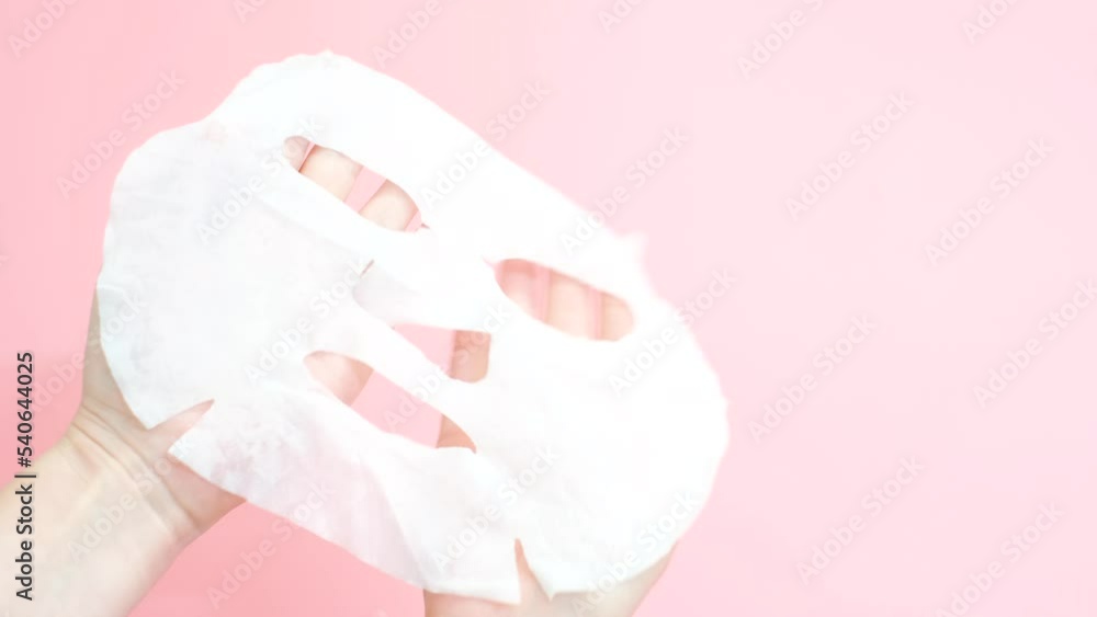 Female hand holding a white sheet of moisturizing mask close-up on a pink background. Female hand holding a white sheet of moisturizing mask close-up on a pink background.