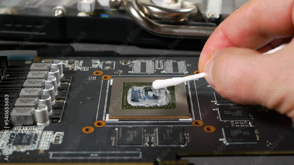 A computer technician removes dried thermal paste from a video card chip using a cotton swab, close-up, selective focus. A computer technician removes dried thermal paste from a video card chip using a cotton swab, close-up, selective focus.