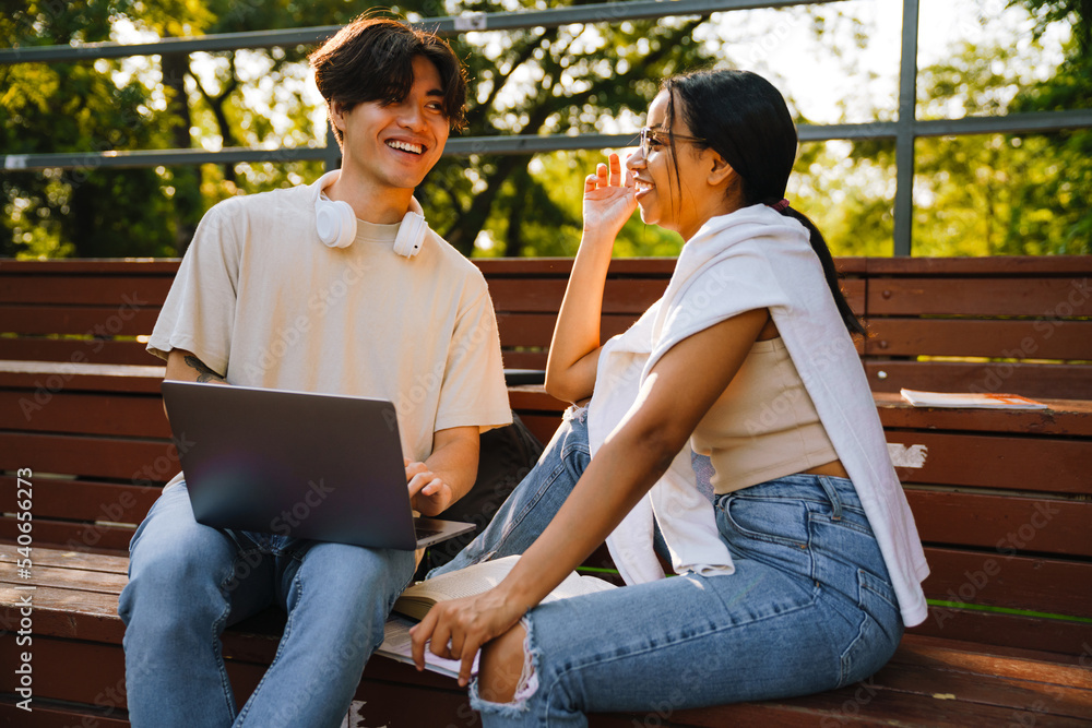 Fototapeta premium Two multiracial friends laughing while using laptop together in skate park