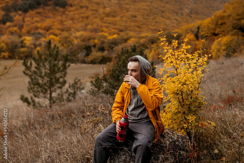 young man in a yellow jacket drinking hot tea from a thermos at the autumn forest