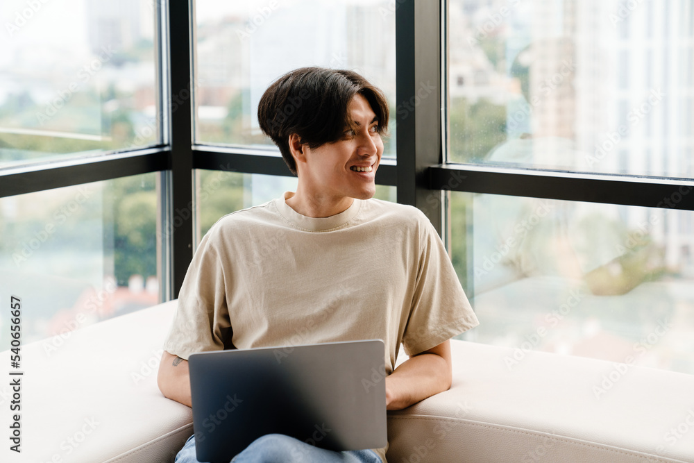 Asian young man using laptop computer while sitting by window at home Asian young man using laptop computer while sitting by window at home