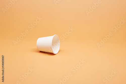 Disposable white single one recyclable cardboard paper cup knocked over the surface isolated on the bright solid fond plain sandy beige background