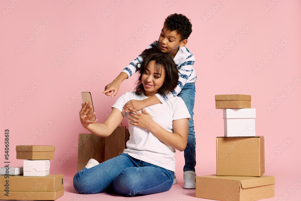 Portrait of woman with her son doing online order on phone, sitting around boxes isolated over pink studio background Portrait of woman with her son doing online order on phone, sitting around boxes isolated over pink studio background