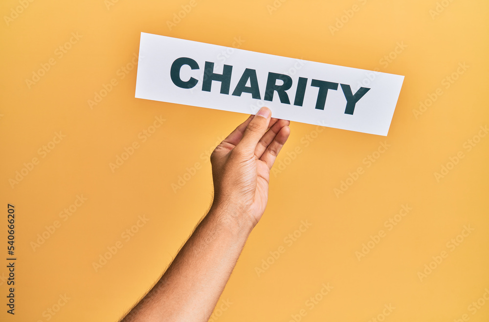 Hand of caucasian man holding paper with charity word over isolated yellow background Hand of caucasian man holding paper with charity word over isolated yellow background