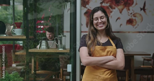 Happy female staff crossing arms smiling at camera in front of coffee shop store wearing yellow apron. A hispanic latin adult girl barista employee of small business restaurant place