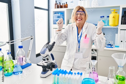 Middle age blonde woman working at scientist laboratory celebrating mad and crazy for success with arms raised and closed eyes screaming excited. winner concept