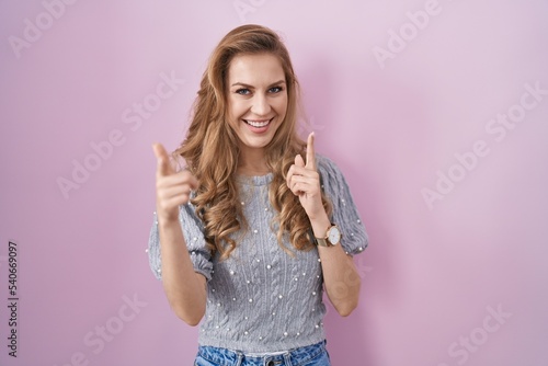 Beautiful blonde woman standing over pink background pointing fingers to camera with happy and funny face. good energy and vibes.