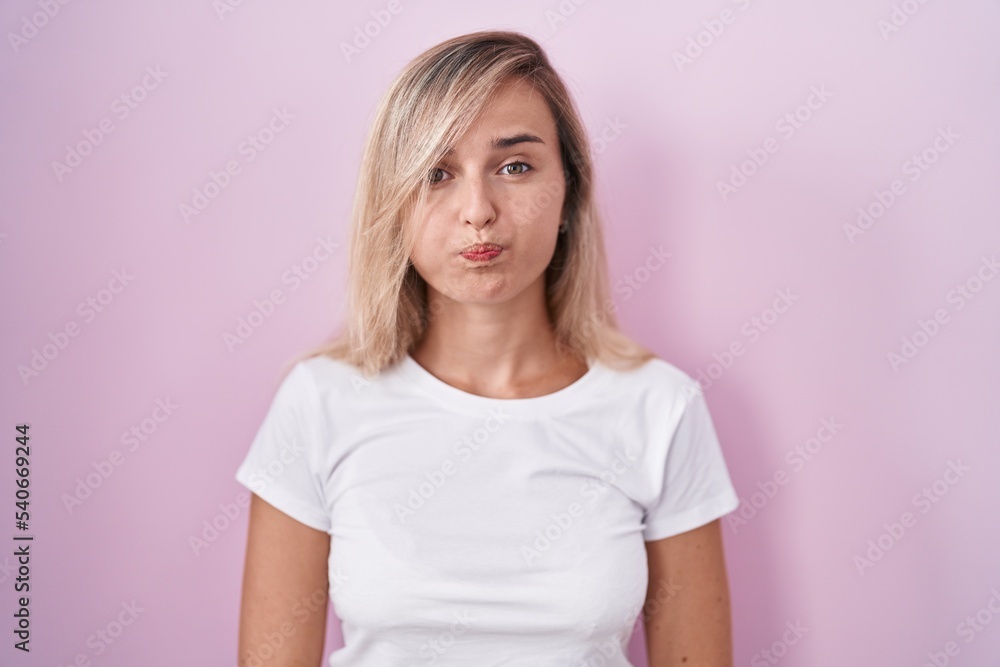Young blonde woman standing over pink background puffing cheeks with funny face. mouth inflated with air, crazy expression. Young blonde woman standing over pink background puffing cheeks with funny face. mouth inflated with air, crazy expression.