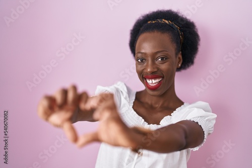African woman with curly hair standing over pink background smiling in love doing heart symbol shape with hands. romantic concept.