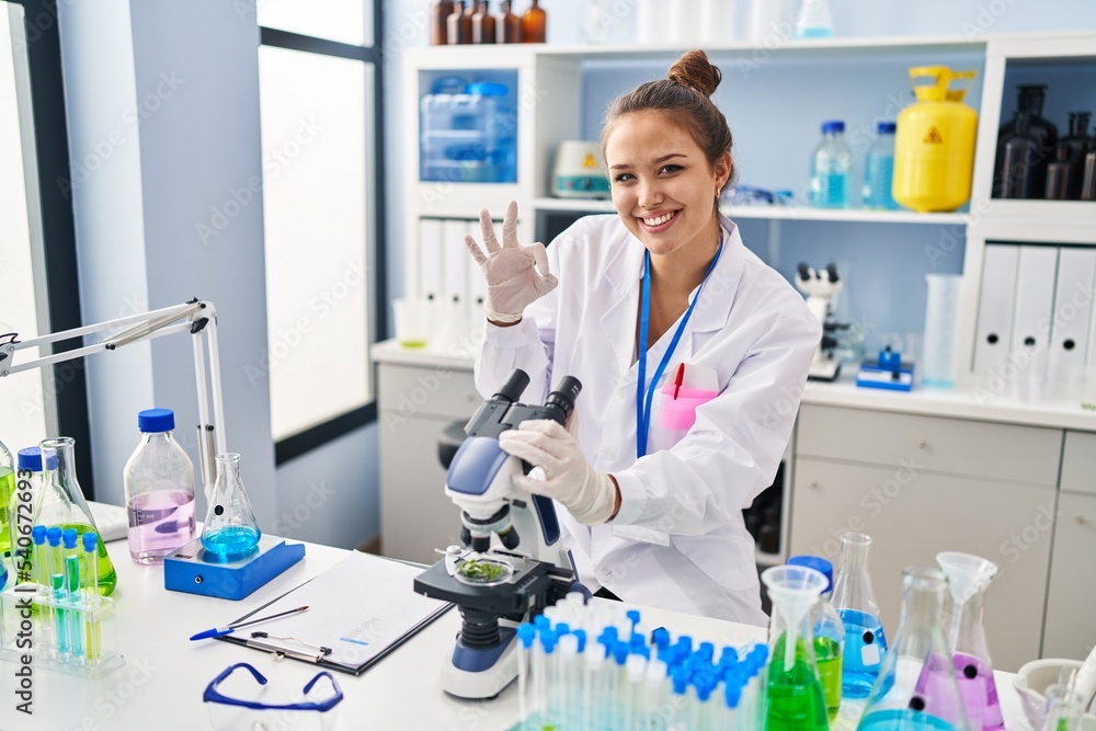 Young hispanic woman working at scientist laboratory doing ok sign with fingers, smiling friendly gesturing excellent symbol Young hispanic woman working at scientist laboratory doing ok sign with fingers, smiling friendly gesturing excellent symbol