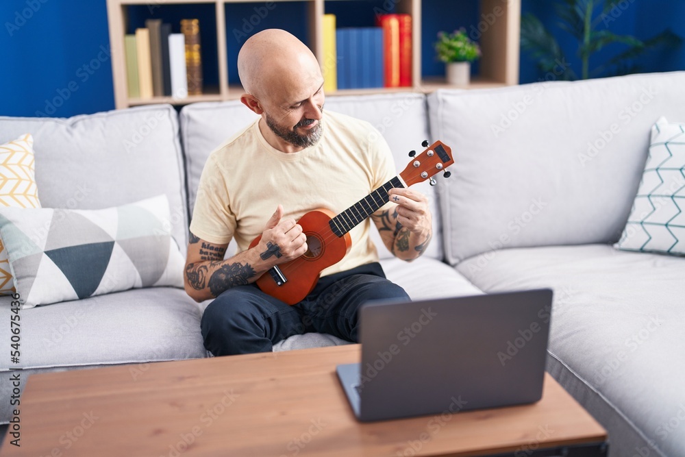 Young bald man having online ukulele class sitting on sofa at home