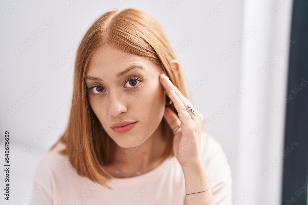 Young redhead woman touching face sitting on bed at bedroom Young redhead woman touching face sitting on bed at bedroom