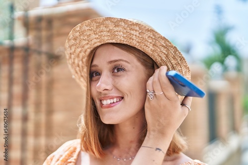 Young redhead woman tourist wearing summer hat listening audio message by smartphone at street