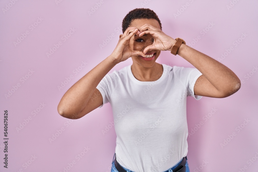 Beautiful african american woman standing over pink background doing heart shape with hand and fingers smiling looking through sign Beautiful african american woman standing over pink background doing heart shape with hand and fingers smiling looking through sign
