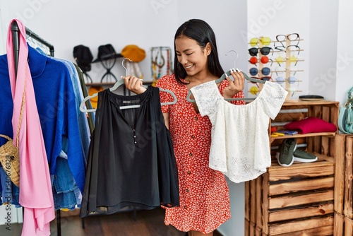 Young latin woman smiling confident holding clothes at clothing store