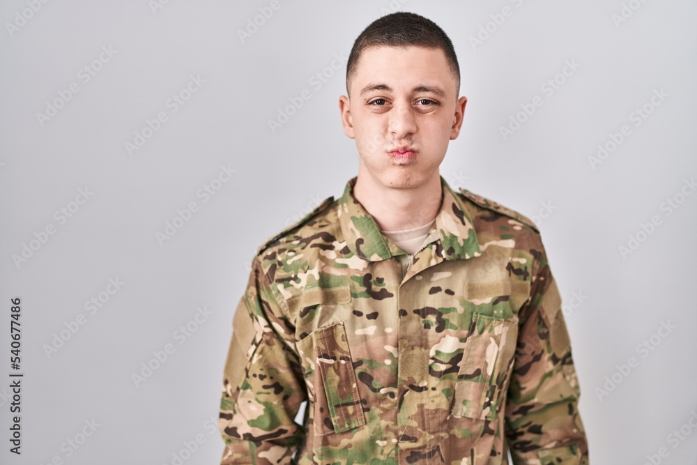 Young man wearing camouflage army uniform puffing cheeks with funny face. mouth inflated with air, crazy expression.
