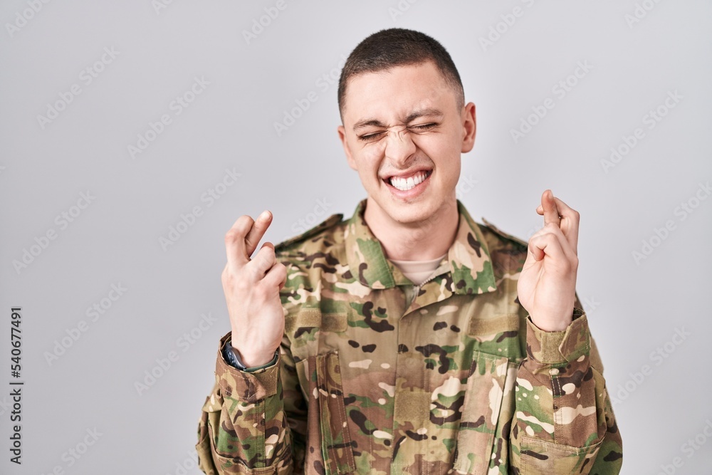 Young man wearing camouflage army uniform gesturing finger crossed smiling with hope and eyes closed. luck and superstitious concept. Young man wearing camouflage army uniform gesturing finger crossed smiling with hope and eyes closed. luck and superstitious concept.