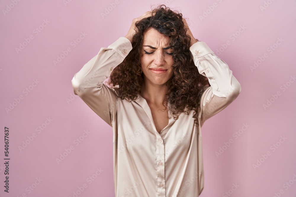 Naklejka premium Hispanic woman with curly hair standing over pink background suffering from headache desperate and stressed because pain and migraine. hands on head.