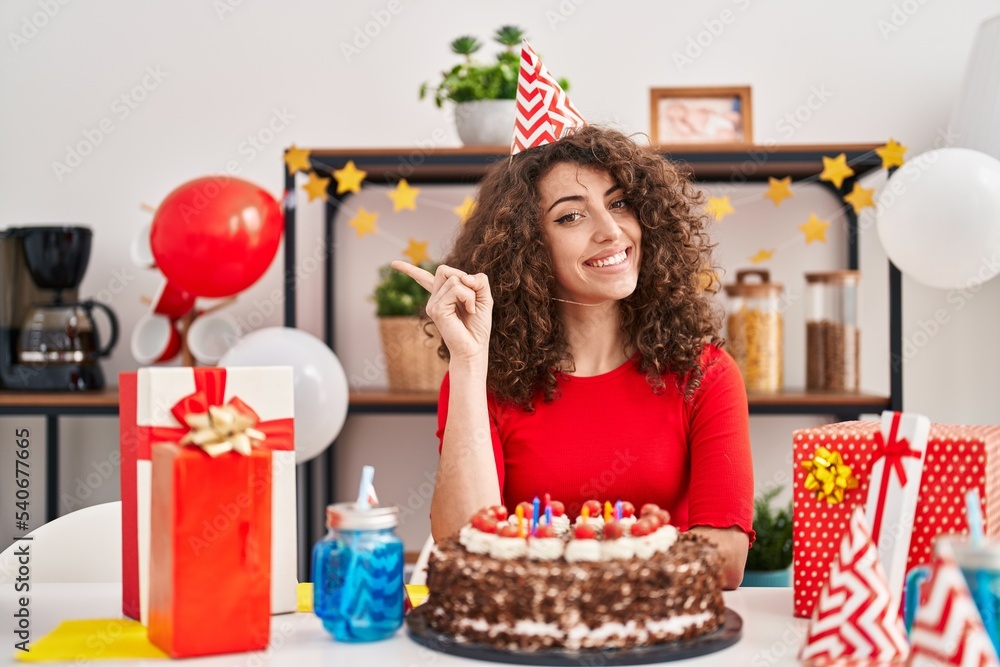 Hispanic woman with curly hair celebrating birthday holding big chocolate cake smiling happy pointing with hand and finger to the side Hispanic woman with curly hair celebrating birthday holding big chocolate cake smiling happy pointing with hand and finger to the side