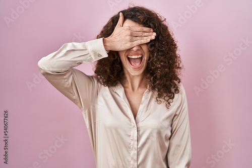 Hispanic woman with curly hair standing over pink background smiling and laughing with hand on face covering eyes for surprise. blind concept. © Krakenimages.com