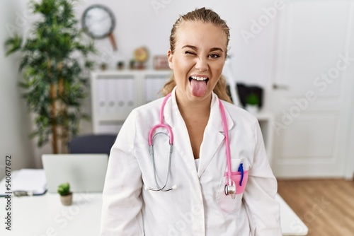 Young caucasian woman wearing doctor uniform and stethoscope at the clinic sticking tongue out happy with funny expression. emotion concept.