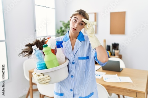 Young blonde woman wearing cleaner uniform holding cleaning products worried and stressed about a problem with hand on forehead  nervous and anxious for crisis