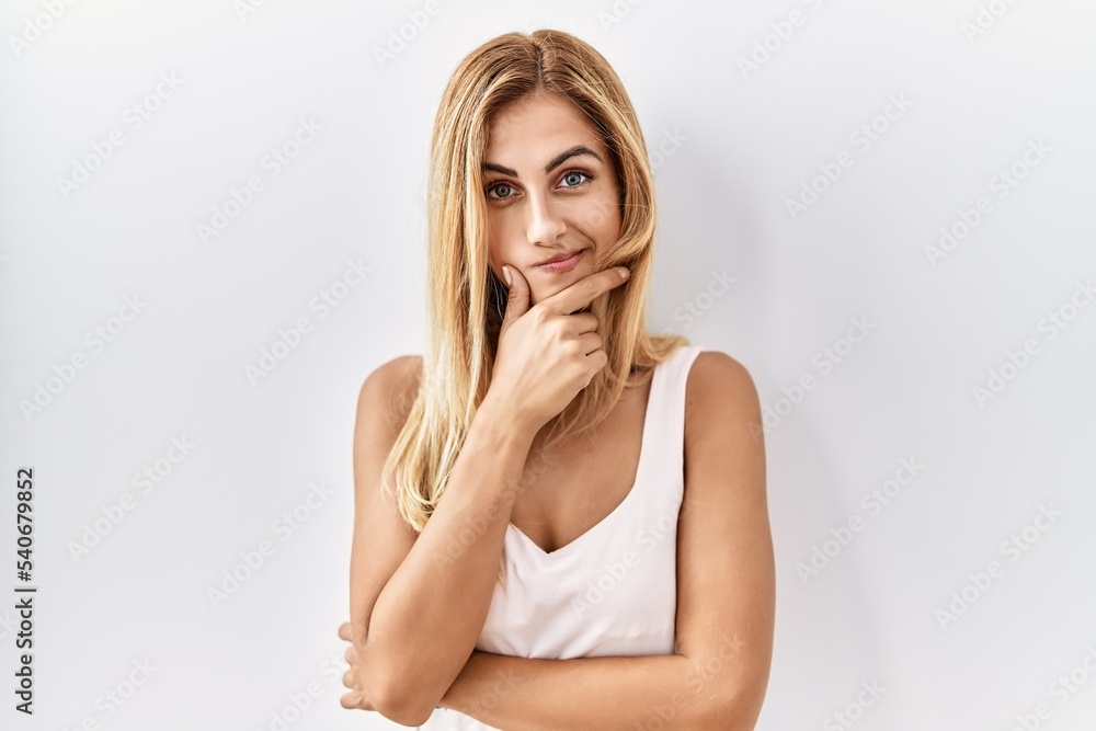 Blonde beautiful young woman standing over white isolated background looking confident at the camera with smile with crossed arms and hand raised on chin. thinking positive. Blonde beautiful young woman standing over white isolated background looking confident at the camera with smile with crossed arms and hand raised on chin. thinking positive.